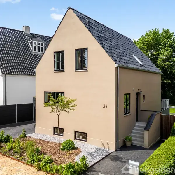 Et beige hus med sort tag står i en forstadsgade omgivet af grønne træer og naboer. Huset har en udvendig trappe og er nummereret med "23" ved hoveddøren.
