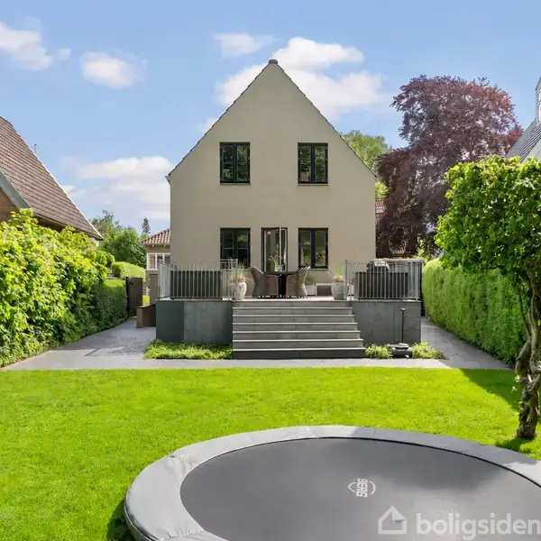 Et moderne hus står stille i en velplejet have med en trampolin i forgrunden. Græsplænen omgives af høje hække, og huset har en terrasse med trapper.
