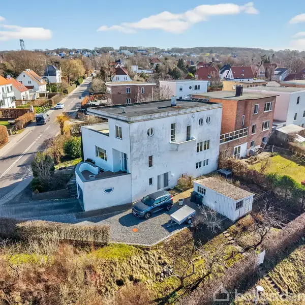 En hvid bygning med flade tage står stille ved en vej i et beboelseskvarter. En bil er parkeret foran. Omkring er andre huse og træer.