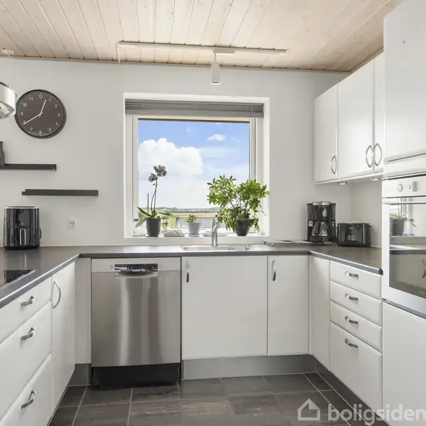 Køkken med hvide skabe og moderne apparater. Over vasken er et vindue med udsigt til en blå himmel. Planter står ved vinduet, og en vægklokke hænger ved siden af.