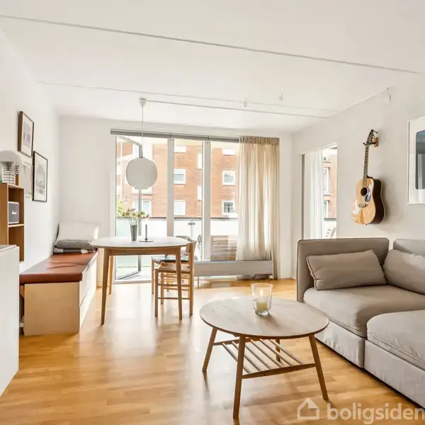 En lys stue med en grå hjørnesofa står ved et rundt sofabord. Til højre hænger en guitar på væggen. Et spisebord med stole ved glasskydedøre.