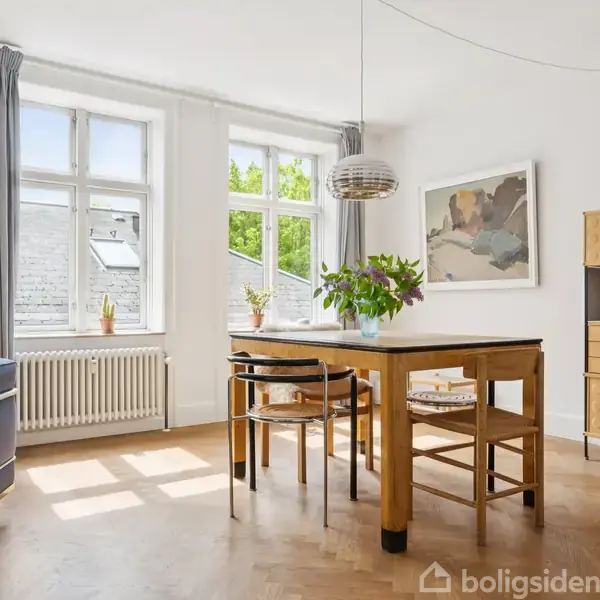 Et lyst spiseområde med et træbord omgivet af stole. En lampe hænger over bordet, og der står en plante på det. Store vinduer lader meget lys ind i rummet.