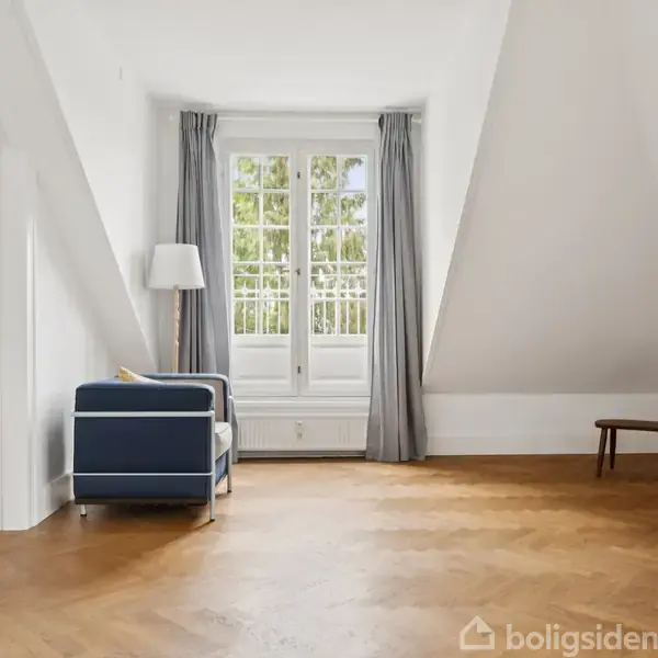 Stue med to blå sofaer, gulvlampe og lille bord med lampe; store vinduer med grå gardiner; trægulv og lyse vægge. Åben forbindelse til tilstødende rum.