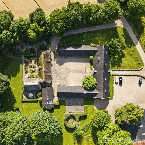 Bygning med gårdsplads ses ovenfra, omgivet af frodig skov og marker. Flere biler holder parkeret langs en grusvej ved siden af gården.