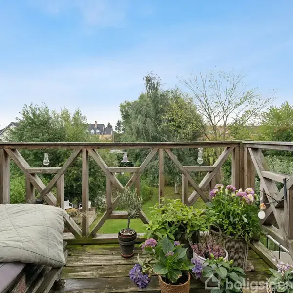 Træterrasse med planter i potter og en sofapude, omkranset af trægelænder med lyspærer. Udsigt over en grøn have med træer i baggrunden under en blå himmel.