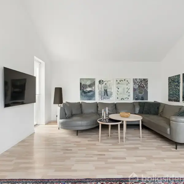 En stor, grå hjørnesofa står i en lys stue. Ved siden af er et lille bord med dekorationer. Væggen har kunstværker, og der er et TV på en anden væg.