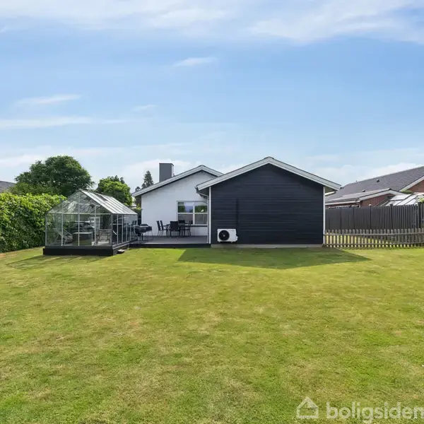 Et sort og hvidt hus står i en velplejet have med en lille gennemsigtig pavillon ved siden af, omgivet af hegn og andre huse i baggrunden.