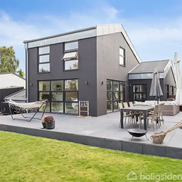 Et moderne, gråt hus med store vinduer. En terrasse har udendørs møbler og en hængekøje. Græsplænen omgiver terrassen. Baghaven er omgivet af buske og træer.