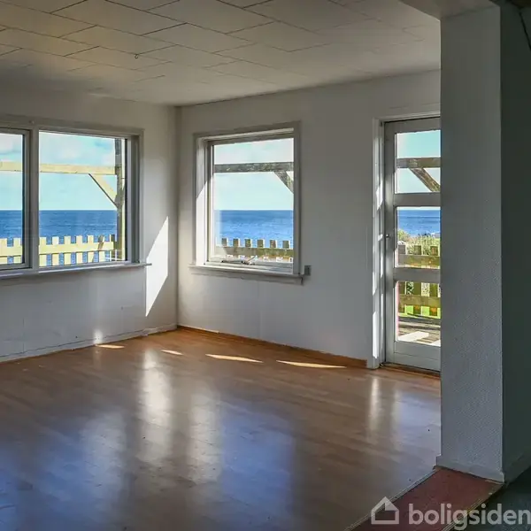 Stue med trægulv, tom for møbler, gennem store vinduer ses havudsigt. Lysindfald gennem sidevinduer og terrassedør. Udenfor en terrasse med rækværk, som er delvist synlig.