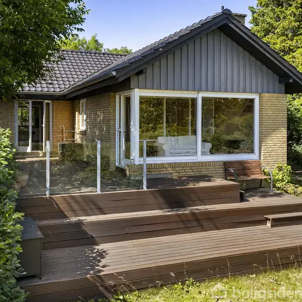 Huset er et murstensbyggeri med store vinduer, der vender ud mod en træterrasse. Terrassen har glasrækværk og er omgivet af frodig have med buske og græsplæne.