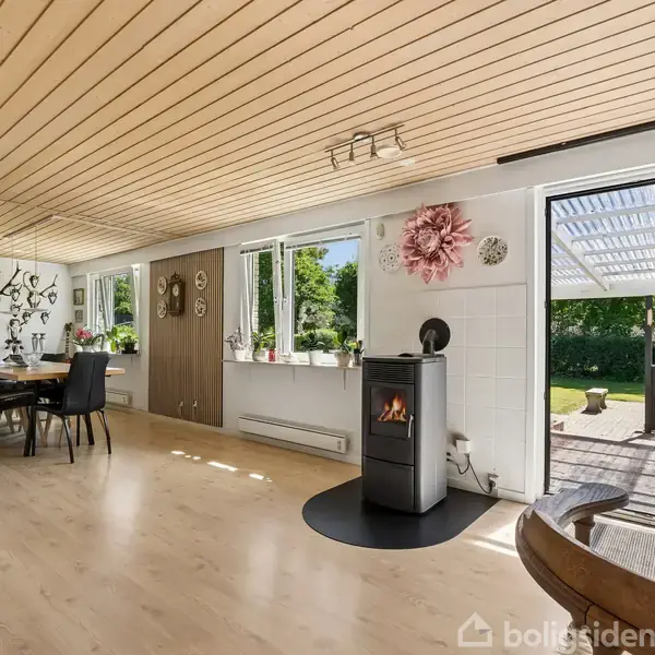 Pejs opvarmer rummeligt køkken-alrum med lyse trægulve. Bord med stole til venstre. Store vinduer og døre åbner mod terrasse og have. Dekorationer på væggen.