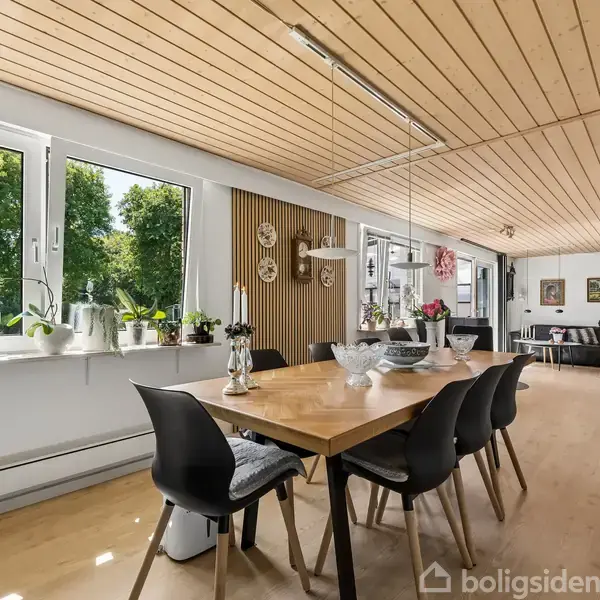 Træbord med sorte stole står i et lyst spisestueområde. Store vinduer med planter og udsigt til grønne træer. Lys trælukkede lofter skaber en hyggelig atmosfære.