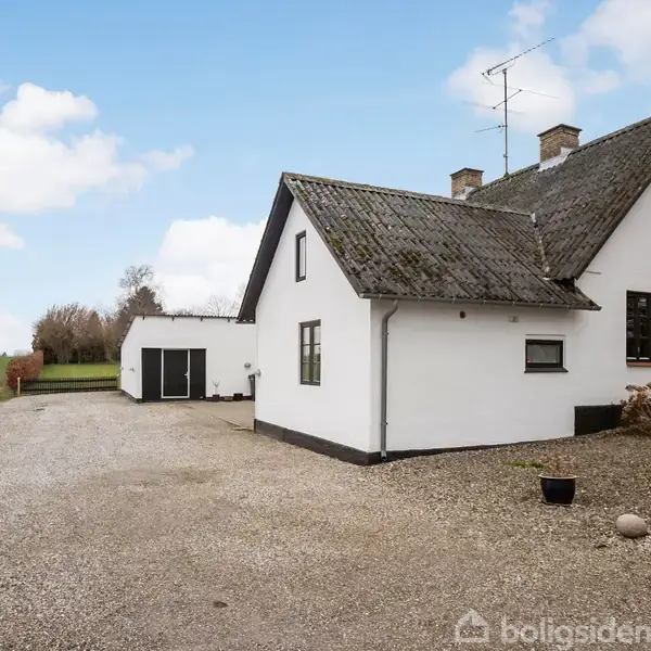 Hvidt hus med skråt tag står stille på en grusvej. Til venstre ses en garagebygning, og i baggrunden er der åbne marker og himmel med lette skyer.