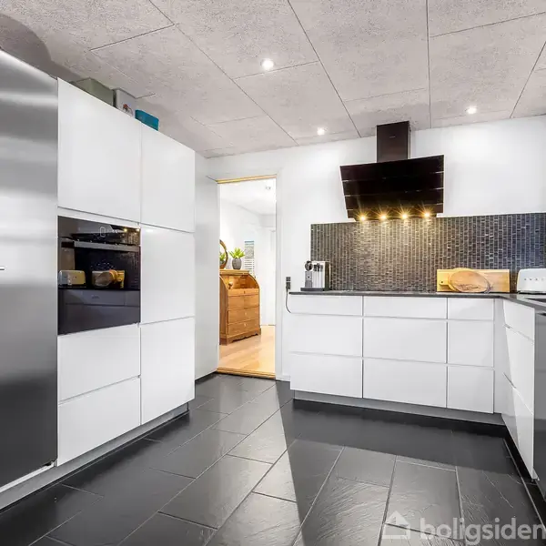 Et moderne køkken med hvide skabe og sort flisegulv. Rustfrit stålkøleskab og indbygningsovn. Sorte fliser bagved håndvasken. Loftets LED-spotlys lyser rummet op. Ingen synlig tekst.