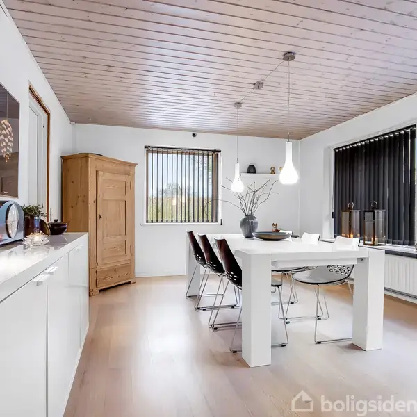 Et lyst spisebord med stole står i et moderne, minimalistisk rum med trægulv og loft. Rummet har store vinduer med lodrette persienner og en hvid skænk med dekorative genstande.