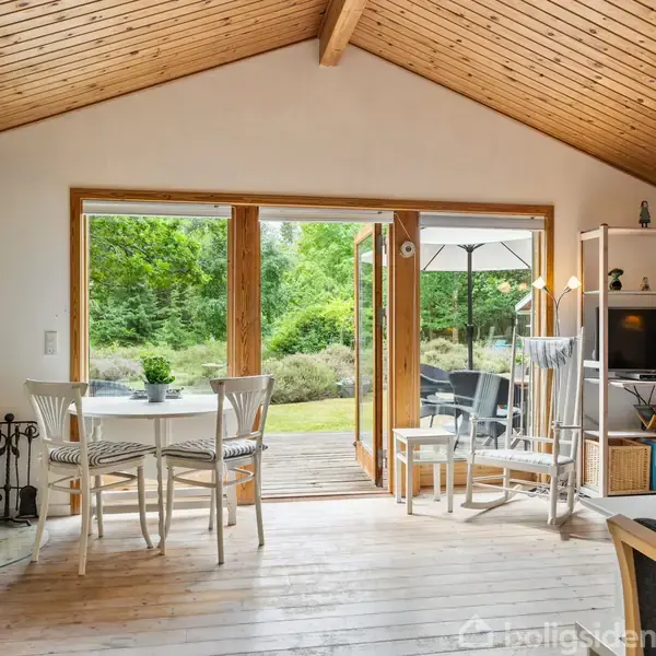 En hyggelig stue med træloft og brændeovn, åbne dobbeltdøre til en terrasse i grønne omgivelser. Et spisebord står centralt, og en hvid reol med TV er til højre.