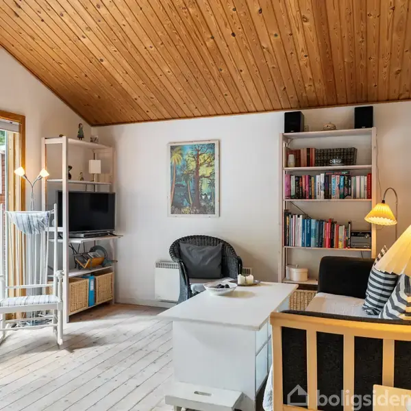 Stue med træloft og lys træindretning. En sofa og lænestol står ved siden af et sofabord. En bogreol og tv står langs væggen. Vindue og terrassedør til venstre.