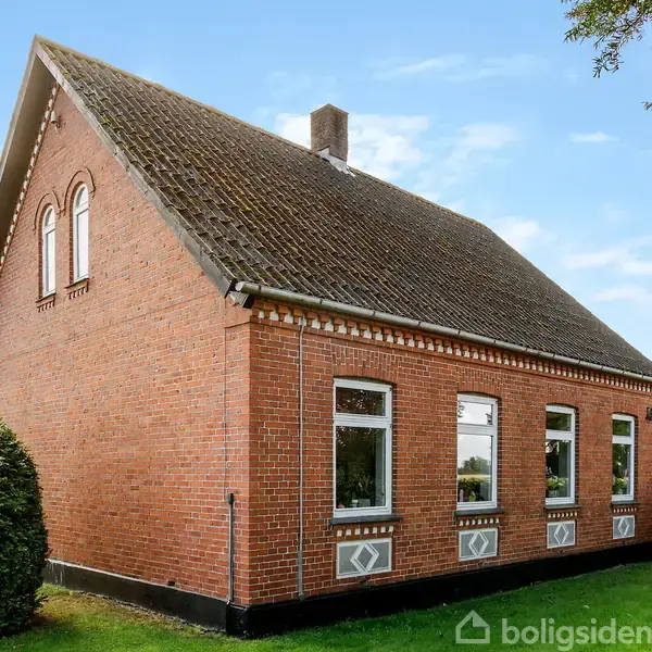 Rødt murstenshus har gavl med skråt mørkt tag, omgivet af grønne buske og træer under en blå himmel. Seks store vinduer langs siden slipper lys ind.