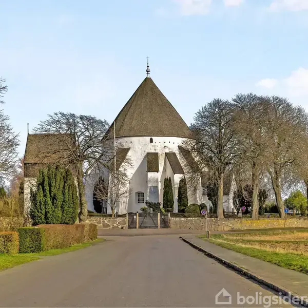 En rund kirke står majestætisk blandt nøgne træer. Kirken har en konisk tagkonstruktion. Vejen foran er omgivet af marker og en velplejet have.