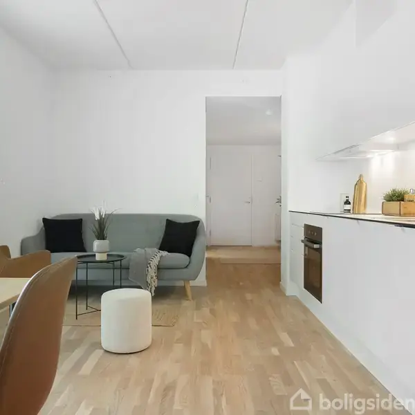 Stue med sofa og sofabord placeret ved siden af et stort vindue, med køkken på højre væg udstyret med ovn og vask, i en moderne og minimalistisk indretning.