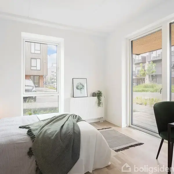 Et soveværelse med en seng dækket af sengelinned og tæppe til venstre, oplyst af en bordlampe. Et skrivebord med stol står ved terrassedøren med udsigt til en have.