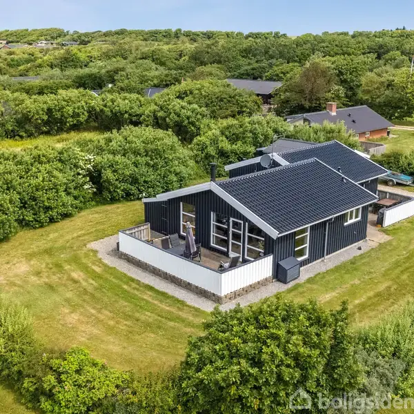 Et sortmalet sommerhus med terrasse ligger omgivet af frodig grøn vegetation og græsområder; huset har store vinduer og en indhegnet grund i en rolig, landskabelig setting.