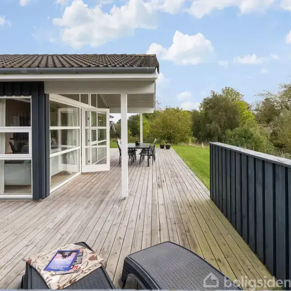 Sortmalet hus med store vinduer står på en træterrasse, omgivet af grønne træer og buske. Der ligger en åben bog på en sort stol, som skaber en hyggelig atmosfære.