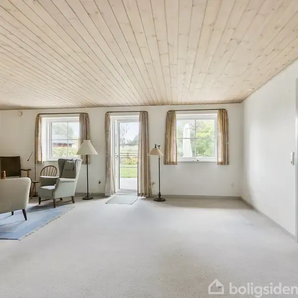 Stue med sofagruppe, stående lamper og store vinduer med udsigt til have. Loftet er i lyst træ, og væggene er hvide. Tæppe på gulvet skaber en hyggelig atmosfære.