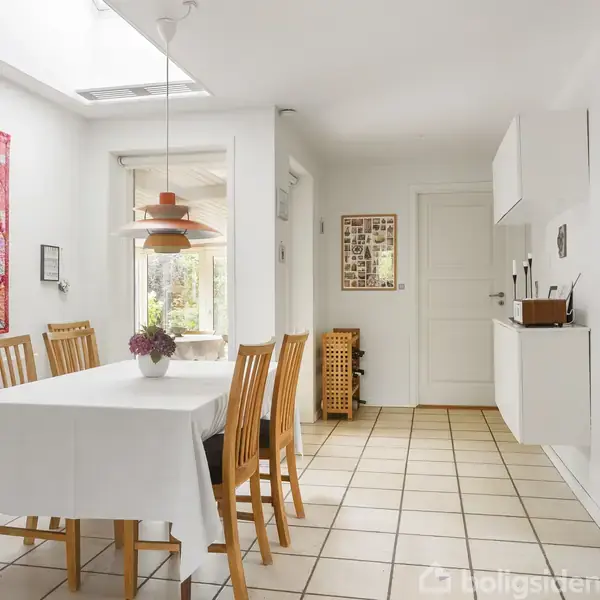 Et spisebord med en hvid dug og træstole står midt i et lyst rum. En farverig vægdekoration hænger til venstre, og en loftslampe hænger over bordet.