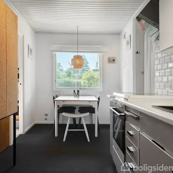 Køkken med hvidt inventar. Et spisebord med to stole står ved et vindue, der giver udsigt til grønne træer. En enkel lampe hænger over bordet.