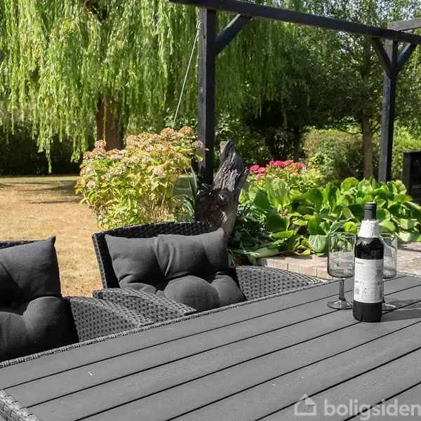 Sorte havemøbler står på en terrasse med et bord, hvorpå der er et glas og en flaske. I baggrunden ses en frodig have med træer og buske.