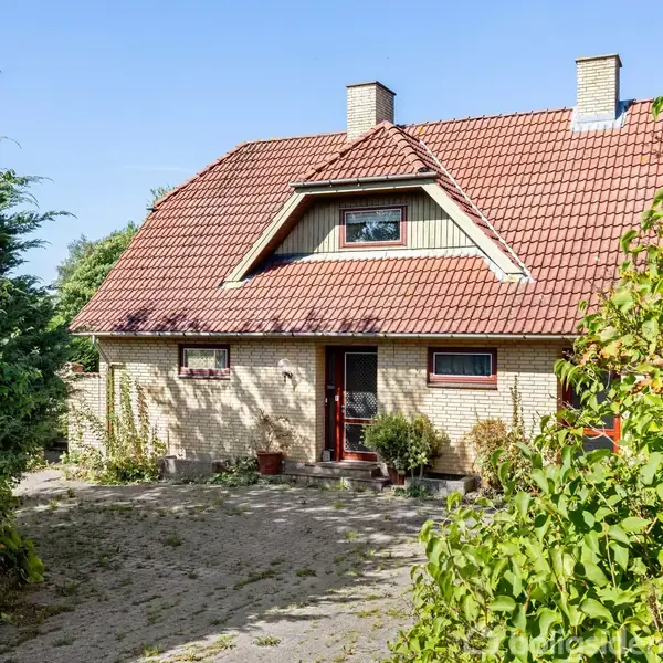 Huset med rødt tegltag har en hoveddør omgivet af planter i krukker. Det ligger i en frodig have med træer og buske og har et klassisk design.
