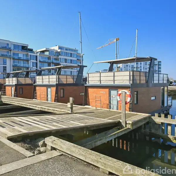 Husbåde ligger til kaj ved en træbro i en havn. I baggrunden ses moderne lejlighedsbygninger under en klar blå himmel. Tekst på båd: "SAUNA".