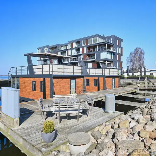 Husbåd med træfacade ligger ved kajen, omgivet af sten og lystbåde. Der er et bord med stole på terrassen. Bagved ses en moderne bygning i flere etager.