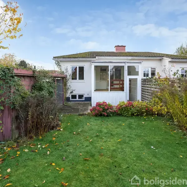 Et hvidt hus med en veranda ses i baggrunden. Foran huset er en grøn græsplæne omgivet af farverige buske. Til venstre står et lille brunt skur.