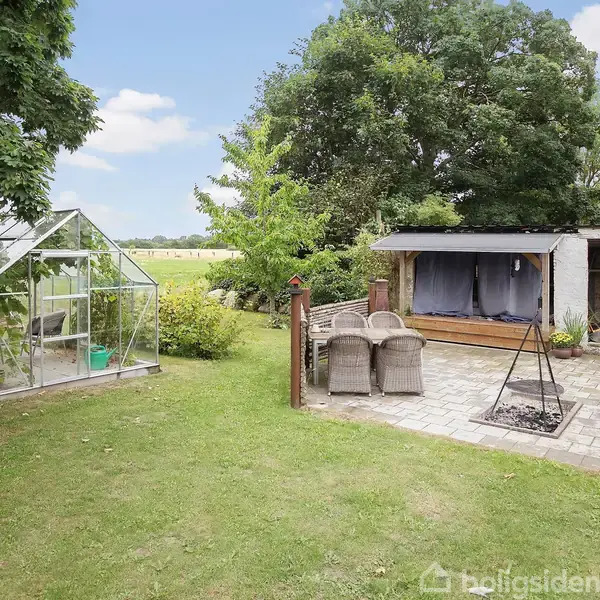 Drivhus står stille i solrig have ved siden af brolagt terrasse med stole og ildsted. Skuret i baggrunden er omkranset af høje træer og græsplæne.