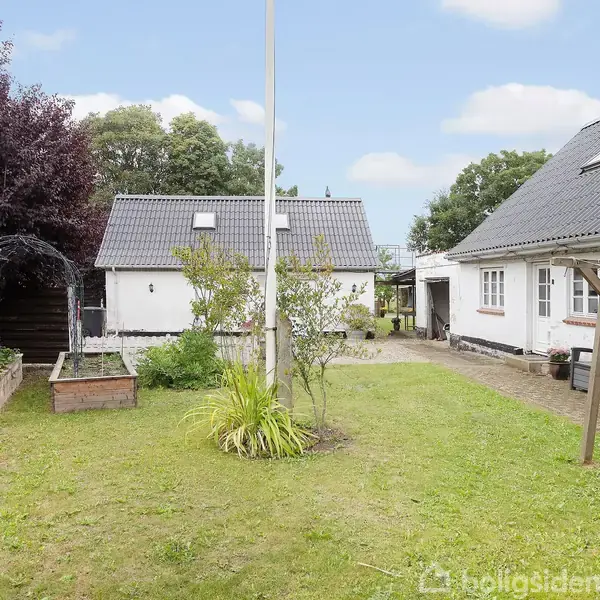 Huset har hvide vægge og mørke tage, omgivet af en velplejet græsplæne og højbede. En flagstang står i midten, og omgivelserne er frodige med træer og buske.