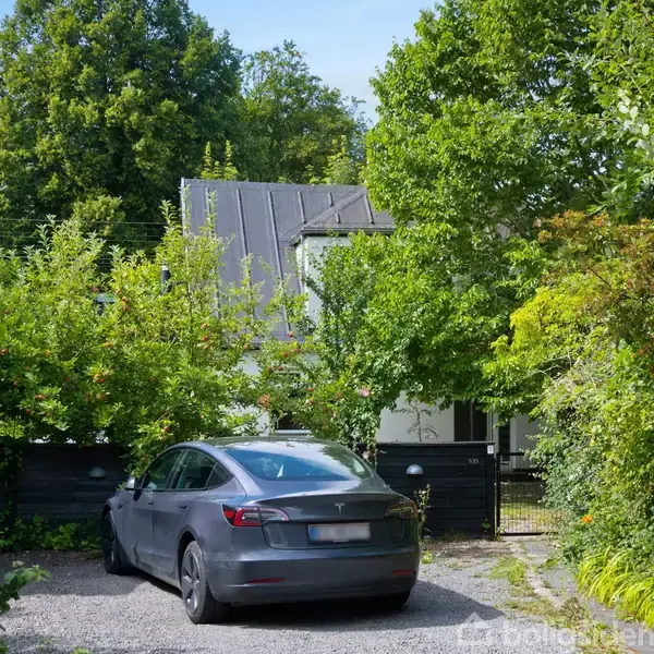 Bil parkeret på grusvej, omkranset af frodige grønne træer og buske. I baggrunden ses et hus med en sort tag og venstredrejet skorsten. Solrigt vejr.