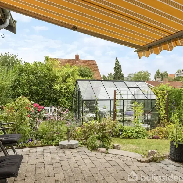 En terrasse med tre sorte stole står under en gul-stribet markise. I baggrunden ses et drivhus omgivet af frodig have med blomster og buske.