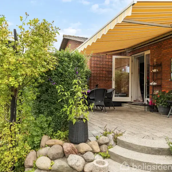 Terrasse med havemøbler under en gulstribet markise, omgivet af murstensvæg og grøn have. Stentrin fører op til terrassen, dekoreret med planter og buske.