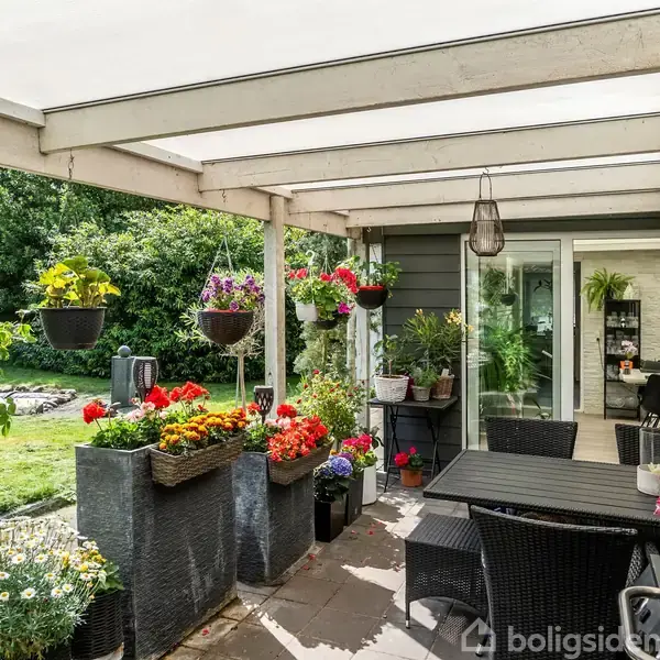 En overdækket terrasse har blomster i krukker og hængende potter. Et sort havebord med stole står centralt. Haven i baggrunden er frodig og grøn.