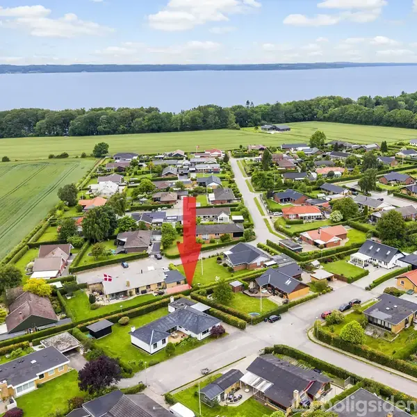 Et parcelhus med en rød pil peger mod det fra oven, omgivet af andre huse i et villakvarter nær en mark med udsigt over vandet i baggrunden.