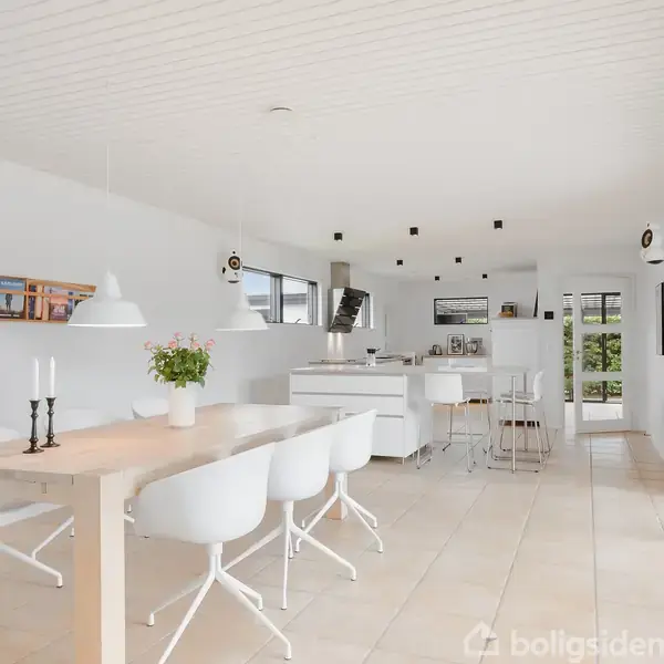 Spisebord med hvide stole står i et lyst køkken-alrum, omgivet af store vinduer og minimalistisk indretning. På væggen hænger hylder med bøger, og der er blomster på bordet.
