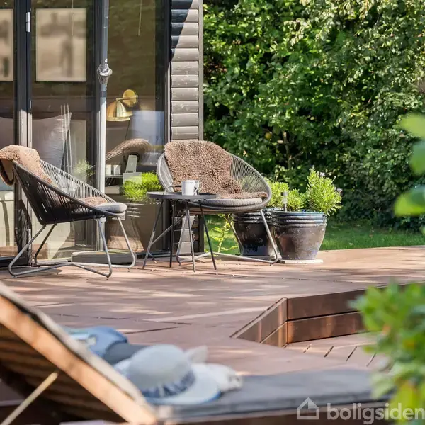 To udendørs stole med pelsindlæg står omkring et lille bord på en træterrasse foran et moderne hus. Omkring terrassen er der frodig grøn beplantning.