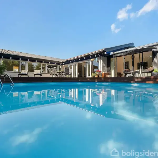 Swimmingpool skinner under blå himmel, omkranset af moderne hus med store vinduer og terrasse. Træer i baggrunden tilføjer naturlig atmosfære.
