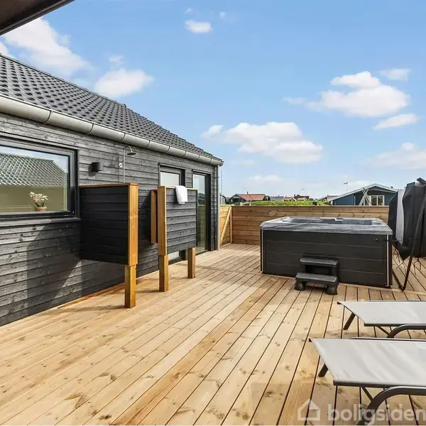 En træterrasse rummer to liggestole og et stort spabad. Baggrunden viser et moderne hus og en himmel med spredte skyer. Tekst: "Modelfoto".