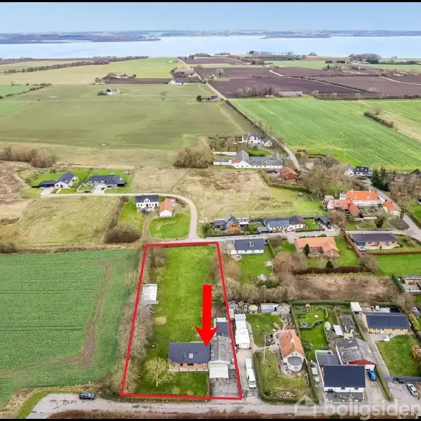 Hus vist fra oven, markeret med en rød ramme og pil. Det ligger i en landlig landsby med marker og spredte huse i nærheden af en kystlinje.