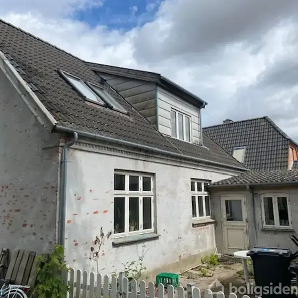 Et hvidt murstenshus med skråt tag og tagvinduer står i en gårdhavelignende baggård. Ved siden af huset ses en skraldespand og en lav haveindhegning. Skyet himmel.