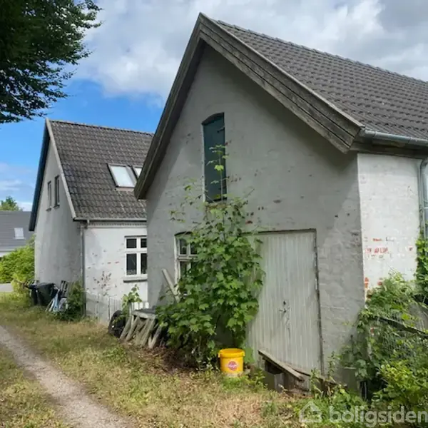 To hvide bygninger med grå tage, omgivet af græs og buske, står ved en smal grusvej. Der er en gul spand og tilfældigt placerede materialer tæt på bygningerne.