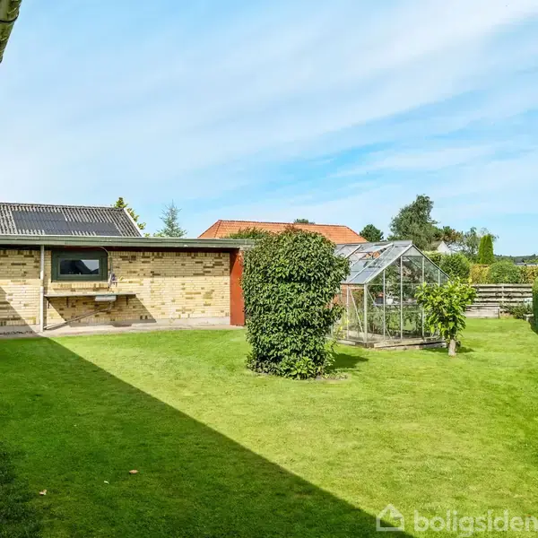 Stenhus står ved siden af en velholdt græsplæne; en glaskasse ses til højre omgivet af hæk og buske; himlen er klar blå med spredte skyer.
